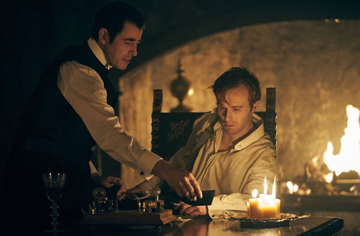 A dark still from the Netflix Series Dracula of a man sitting at a table and a man standing, with a roaring fire behind them.