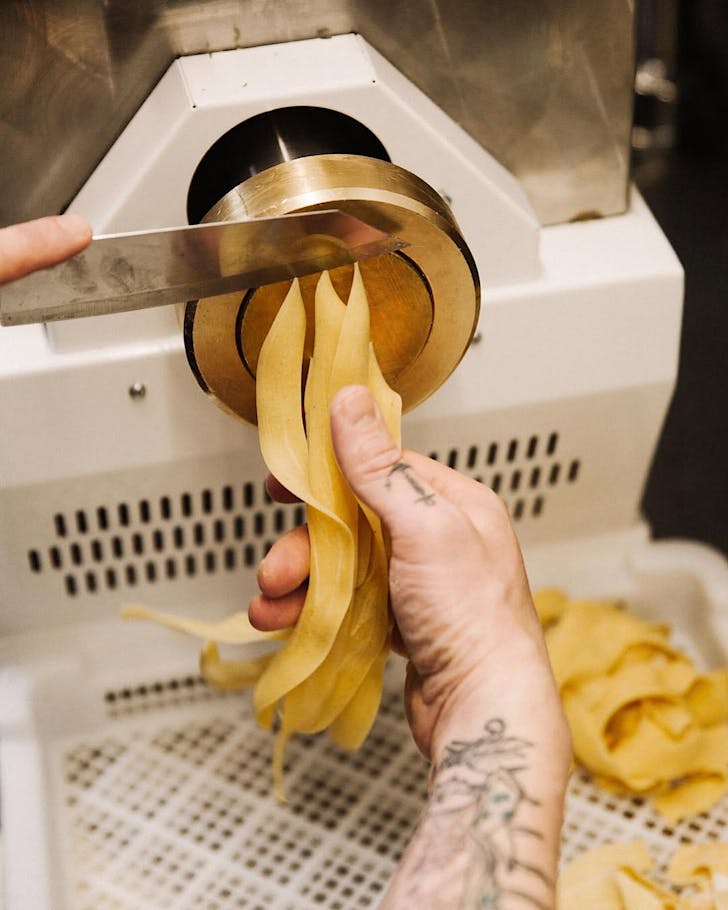 Pasta is extruded from a machine and sliced by somebody with tattooed forearms.