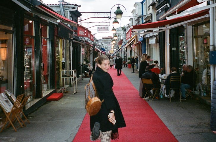 A person in a black coat smiles while standing on a red carpet in a bustling shopping street.