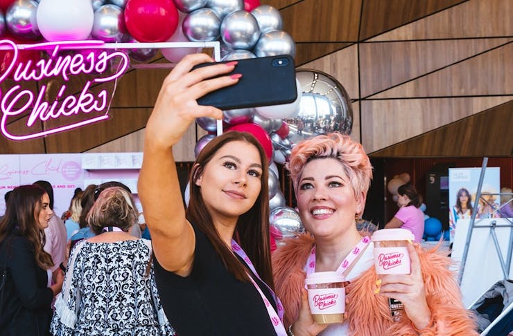Two women take a selfie in front of a pink neon sign.
