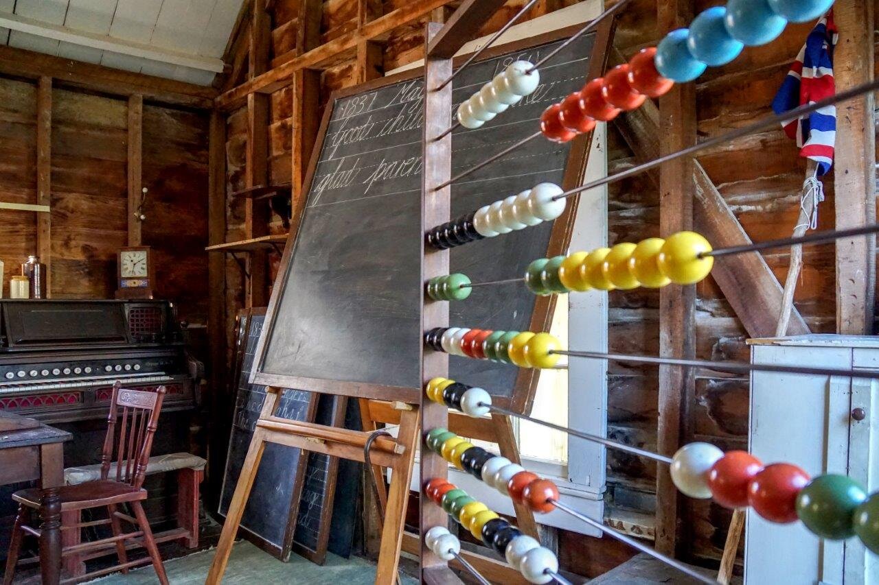 A wooden school house from the 1980s: in the foreground is an abacus with brightly coloured beads, a blackboard with cursive handwriting sits in the mid-ground and at the bag of the room is an organ. 