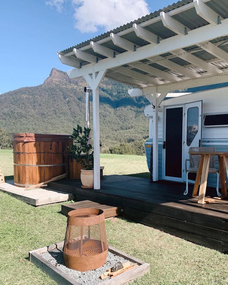 A caravan is parked in a field under blue skies. A hot tub and fire pit are nearby, and a mountain range in the distance.