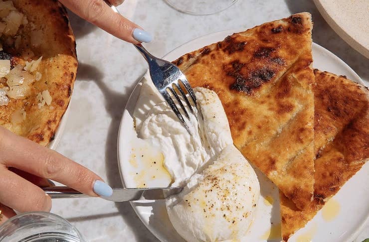 A ball of burrata on a plate with flatbread is pulled apart with cutlery.