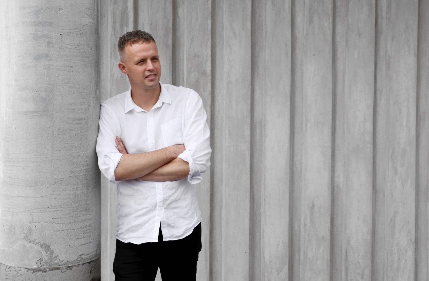 A man stands in front of a grey wall wearing a white shirt and black pants, with his arms crossed.