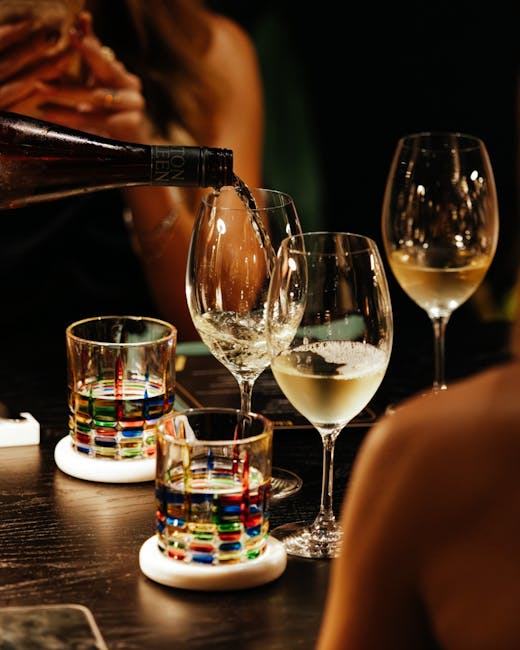 Close-up of white wine being poured into a glass at Avalon Claremont, surrounded by colourful glassware on a dark timber table.