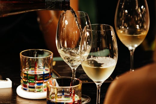 Close-up of white wine being poured into a glass at Avalon Claremont, surrounded by colourful glassware on a dark timber table.