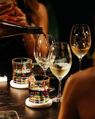 Close-up of white wine being poured into a glass at Avalon Claremont, surrounded by colourful glassware on a dark timber table.