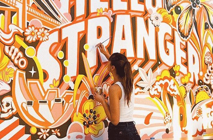 Gemma O'Brien paints a brightly coloured orange mural that reads 'Hey Stranger'.