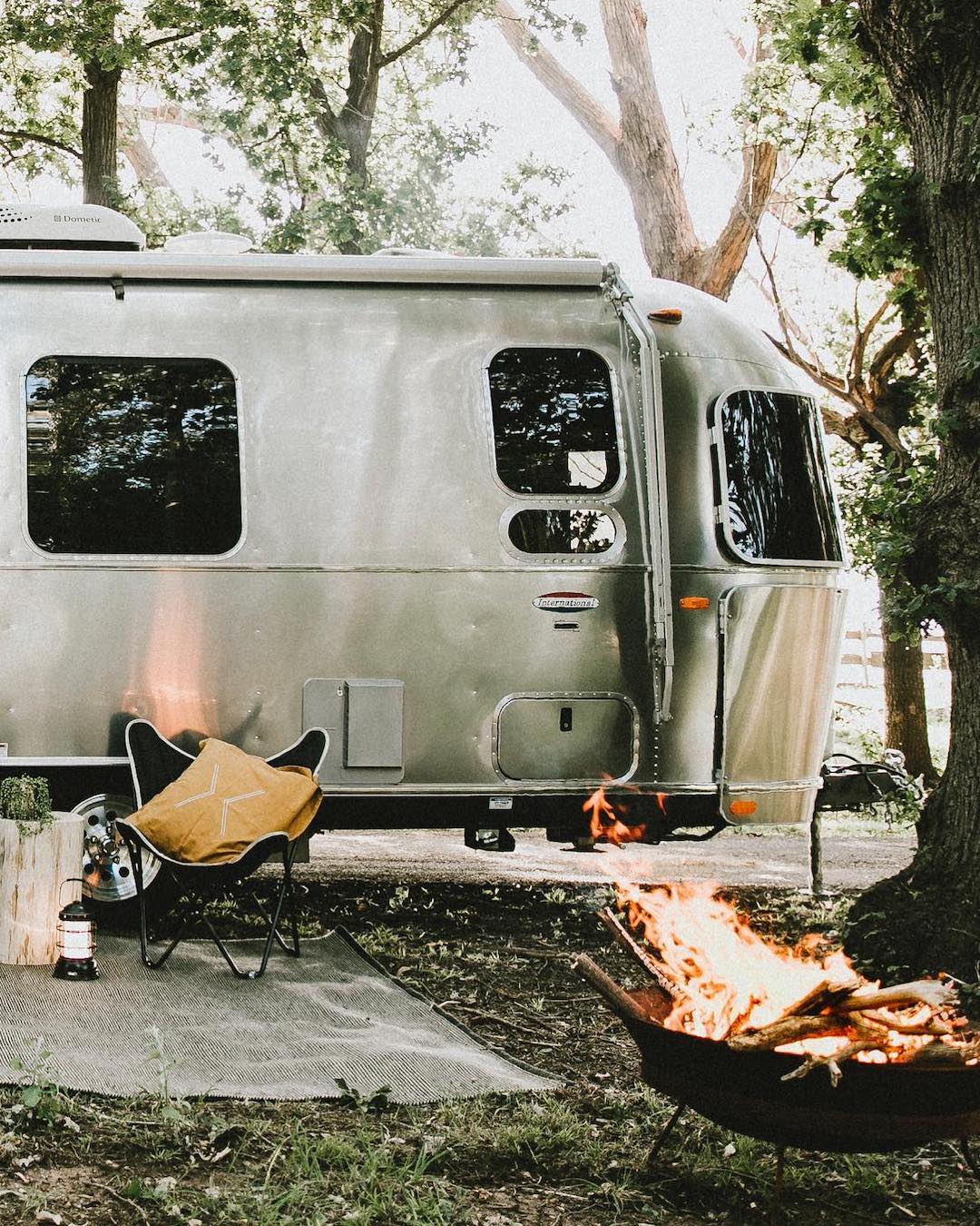 An Airstream is parked under a tree with a campfire burning outside.
