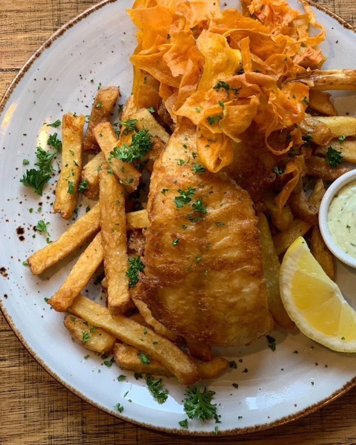 Above view of a battered fish and chips on an elegant plate with aesthetic sweet potato crisps decorating the area, as well as sauce and a lemon. 