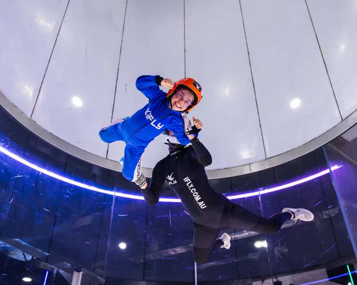 A mid shot of a kid flying in the air wearing an iFly blue suit and helmet with a big smile facing the camera. Behind the child is a trained man in black also flying, and they seem to be positioned in a large tube of sorts that simulates the airflow for skydiving.