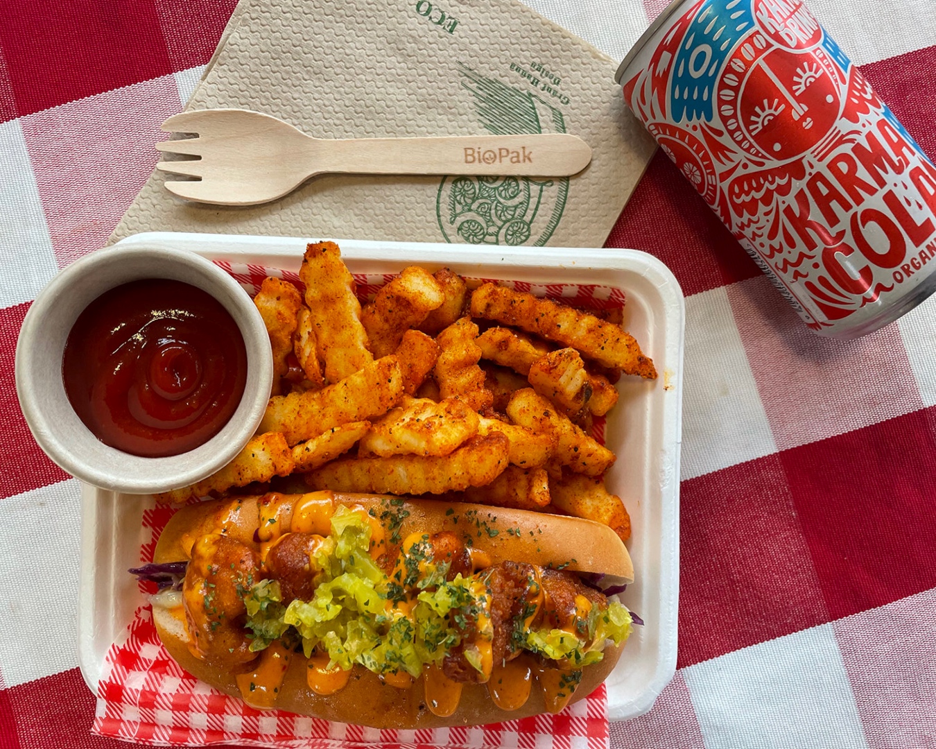A container of seasoned crinkle-cut fries next to a sub roll of fried cauliflower topped with pickles and herbs, a pot of tomato sauce and a can of Karma Cola