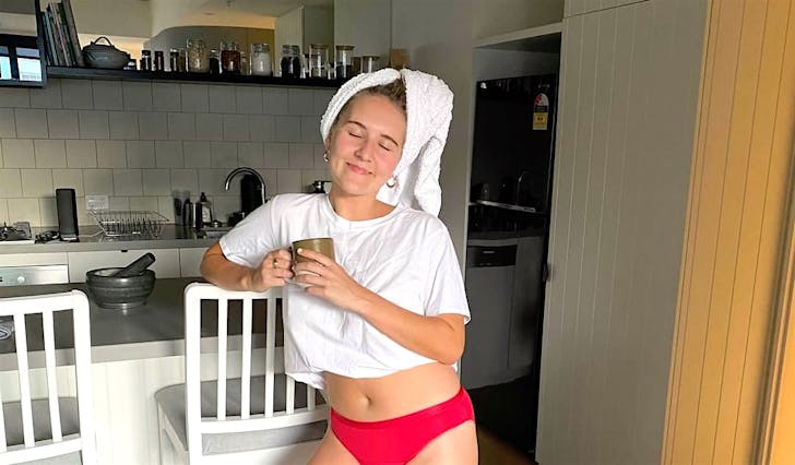 A woman stands in her kitchen holding a cup of coffee in a white t-shirt and red period-proof Modibodi underwear with a towel wrapped around her head, as if she'd just got out of the shower. 