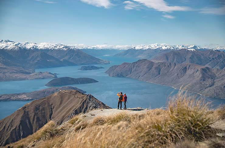 People stand atop Roys Peak Track looking at the amazing white capped mountains and scenery beyond.