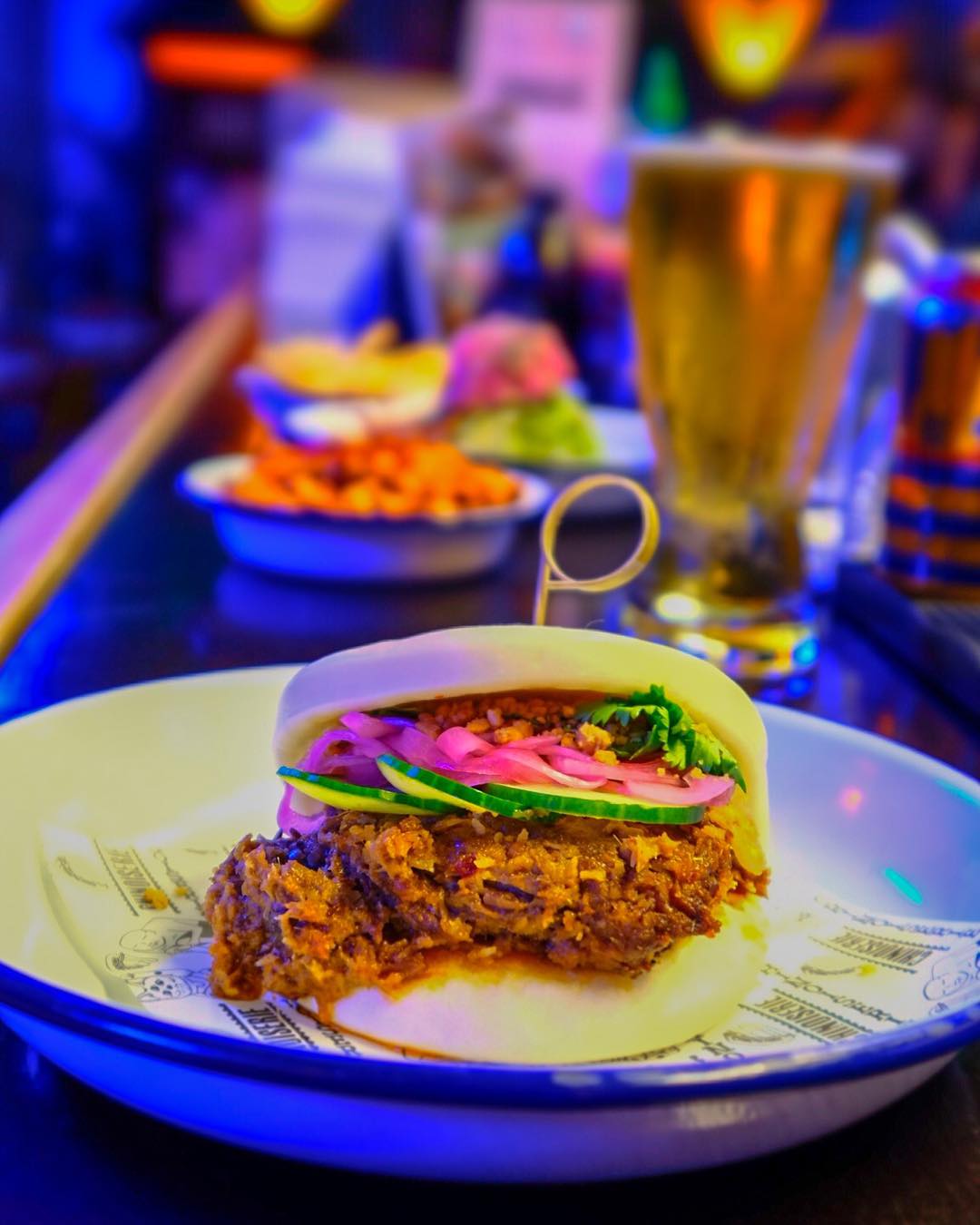 A special fried kumara bao topped with cucumber slices and pickled onion. IN the background are slightly blurry, colourful meals and a pint of beer. 