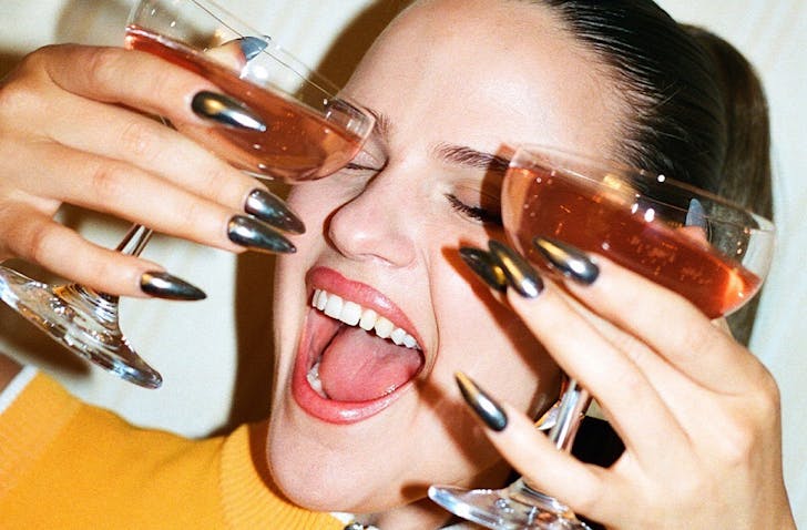 A jovial woman holds two cocktails and has exquisite chrome-style manicured nails. 