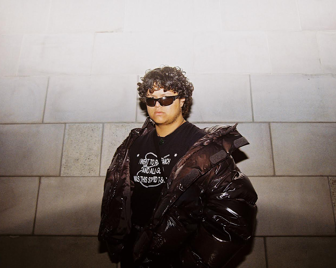 Up and coming DJ ATARANGI stands in front of a block wall wearing a puffer jacket and shades. 