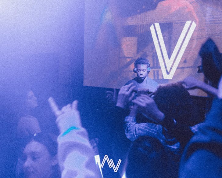 A DJ spins some sick tracks while a crowd loses their minds at AV Club. Everything is bathed in violet light. 