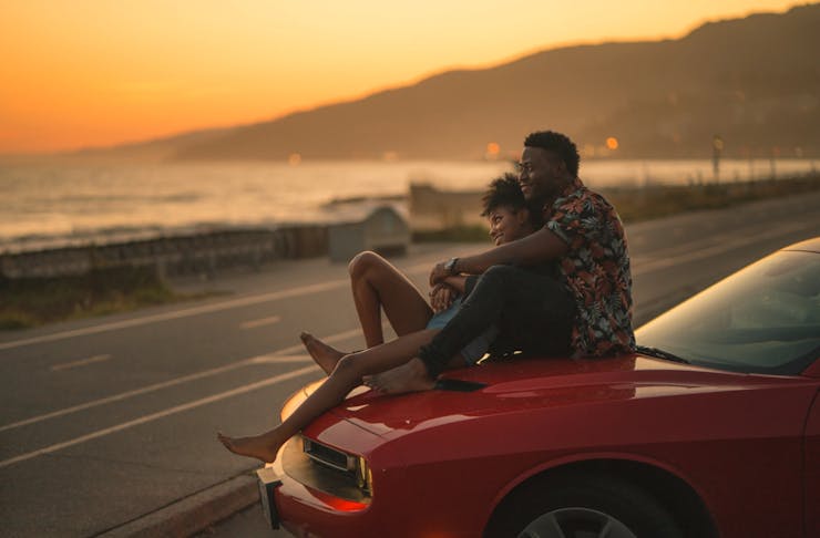 A couple sitting on a red car with a gorgeous sunset in the background. 