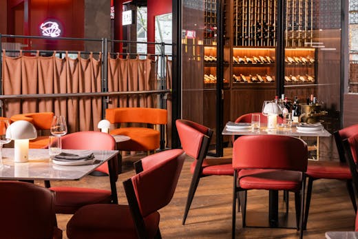 An indoor dining area without any people, featuring wooden floorboards, plush red chairs and a bar in the background.