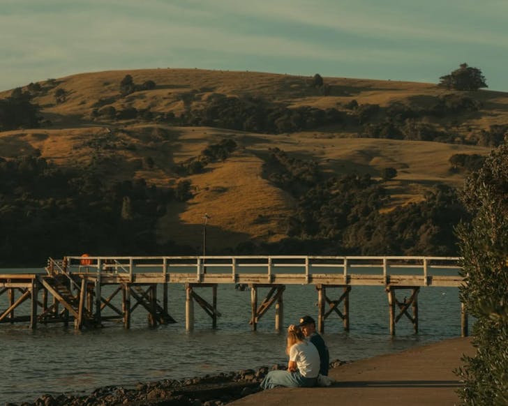 A couple sits by the water at Akaroa, with a wharf and rolling hills in the background. 