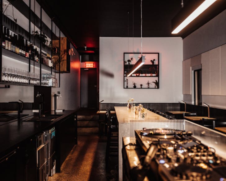 Side view of the inside of Alas bar. There is a vinyl player in the forefront of the image, and a long bench of seating in the background. There is a display bar to the left. The lighting is low and moody. 