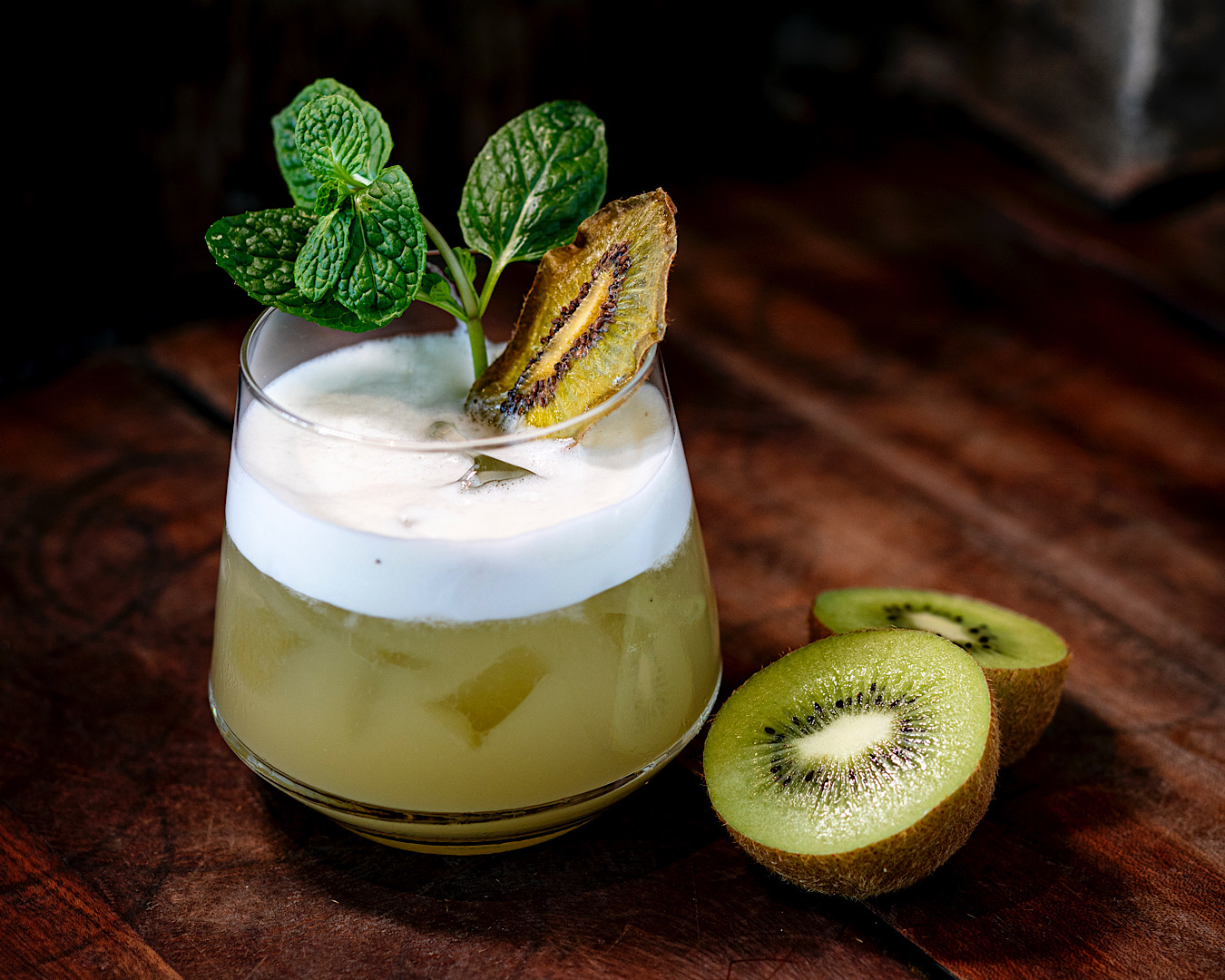 A short glass holds a green cocktail topped with a delightful amount of foam, a freeze-dried slice of kiwifruit and a sprig of mint. A cut kiwifruit half sits beside the glass. 