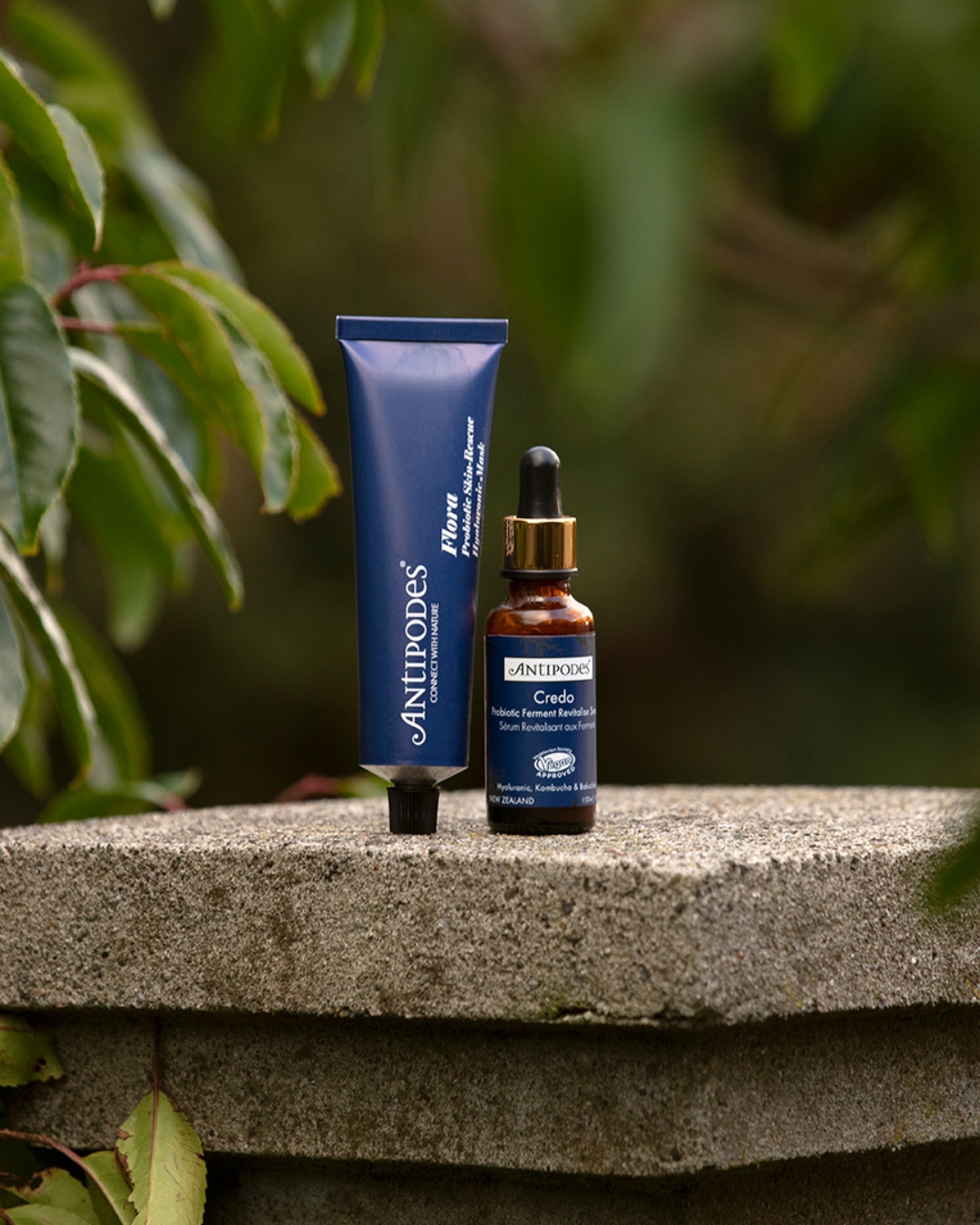 A tube and dropper of Antipodes skincare products with dark blue packaging with green leaves in the background.