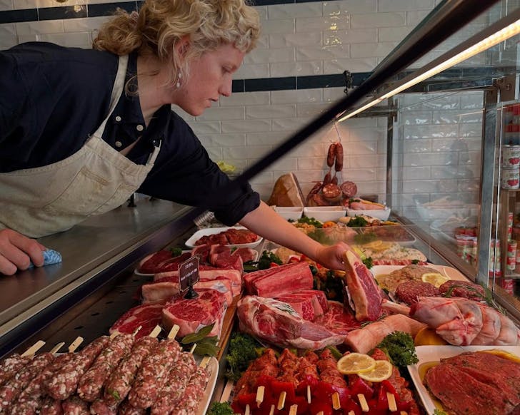 A side view picture of a butcher grabbing a piece of meat inside the display fridge. There are layers and rows of meat inside the fridge.