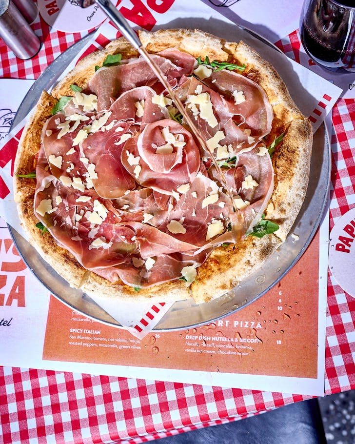 A large prosciutto and parmesan pizza set on top of an Italian checkered tablecloth