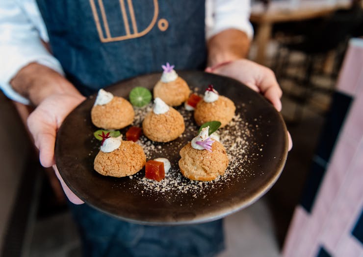a person with a blue apron holding a dish of savoury profiteroles