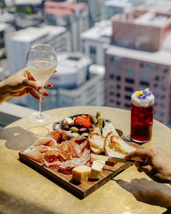 Two people drinking cocktails and tucking into a cheeseboard high up in the air. 