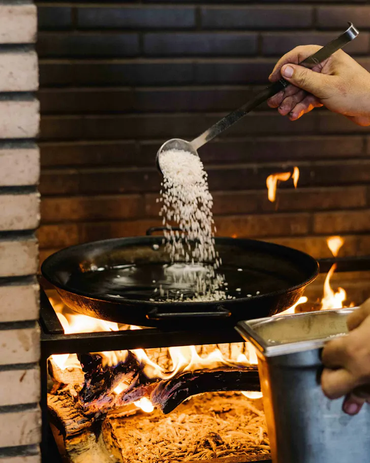 A cook at Barraca tosses rice in a wok over open flames, preparing paella.