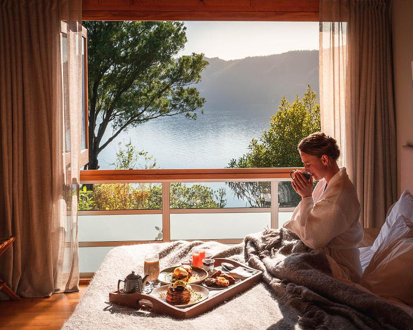 A woman enjoying a coffee and breakfast in bed with views of the Marlborough Sounds outside at Bay of Many Coves Resort.