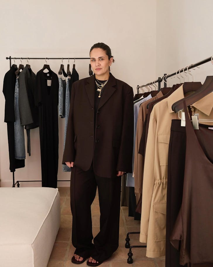 A designer stands amongst racks of elegantly displayed clothing wearing a quality casual blazer and pant set while smiling gently at the camera.