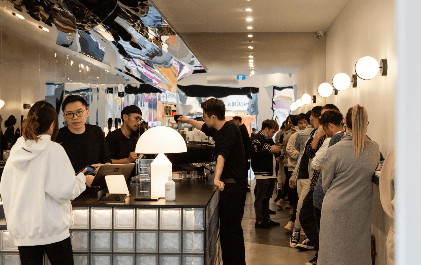 A busy cafe with people standing at a bar and a large overhead mirror. 