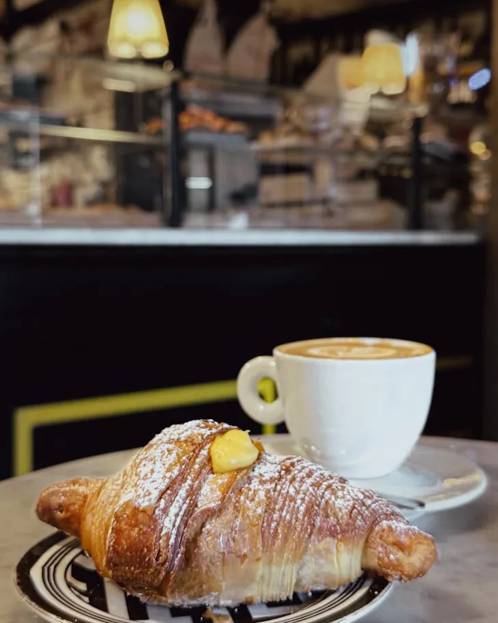 A flaky Italian pastry filled with custard and dusted with icing sugar, served alongside a cappuccino in a cosy Roman cafÃ© setting