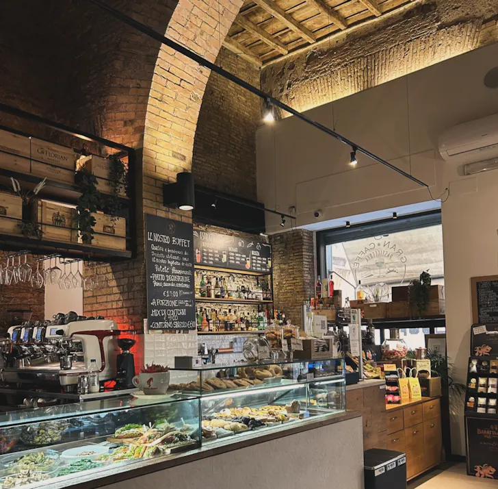 A warm, rustic Italian cafÃ© interior with exposed brick arches, a pastry-filled display counter and a coffee machine in a heritage setting.