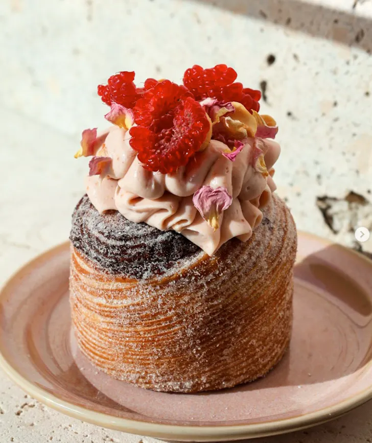 A flaky, sugar-dusted pastry topped with piped cream, fresh raspberries and edible petals on a ceramic plate in soft natural light.