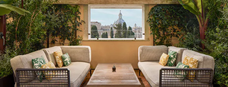 Best rooftop bars in Rome at the Bulgari Hotelâs terrace with an outdoor seating area surrounded by greenery and an open-air window framing Rome