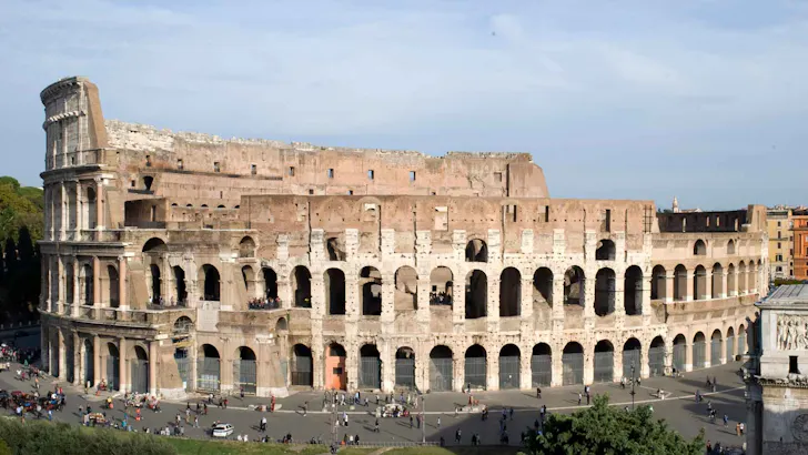 Ancient Roman Colosseum in Rome with its iconic arched exterior, partially ruined upper walls and crowds gathered around the base in a large open piazza