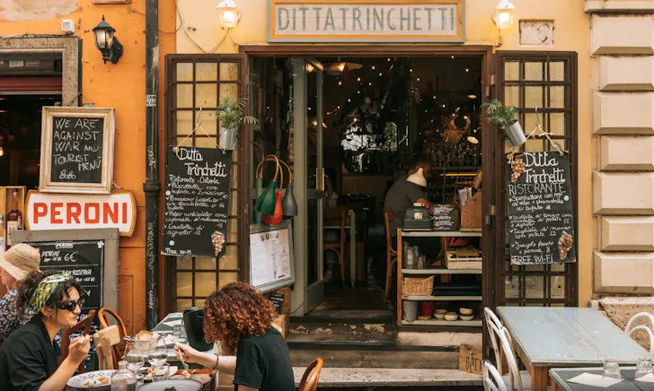Cosy Trastevere street restaurant in Rome with outdoor seating, handwritten menus and locals dining at small tables outside a warm, rustic storefront.