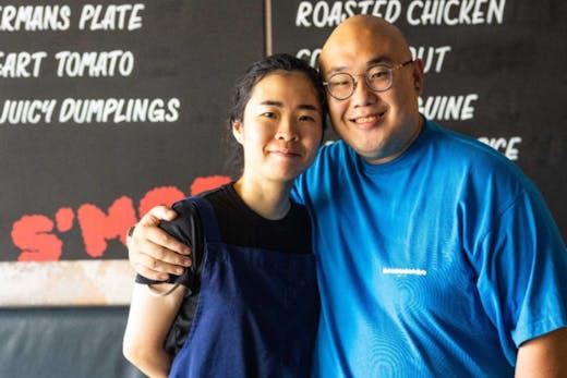 Chef Big Sam Young next to co-chef Grace Chen. Sam Young stands with his arm around Grace Chen's shoulder. Both chefs are smiling welcomingly as they stand in front of a menu on the wall.