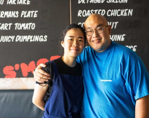 Chef Big Sam Young next to co-chef Grace Chen. Sam Young stands with his arm around Grace Chen's shoulder. Both chefs are smiling welcomingly as they stand in front of a menu on the wall.