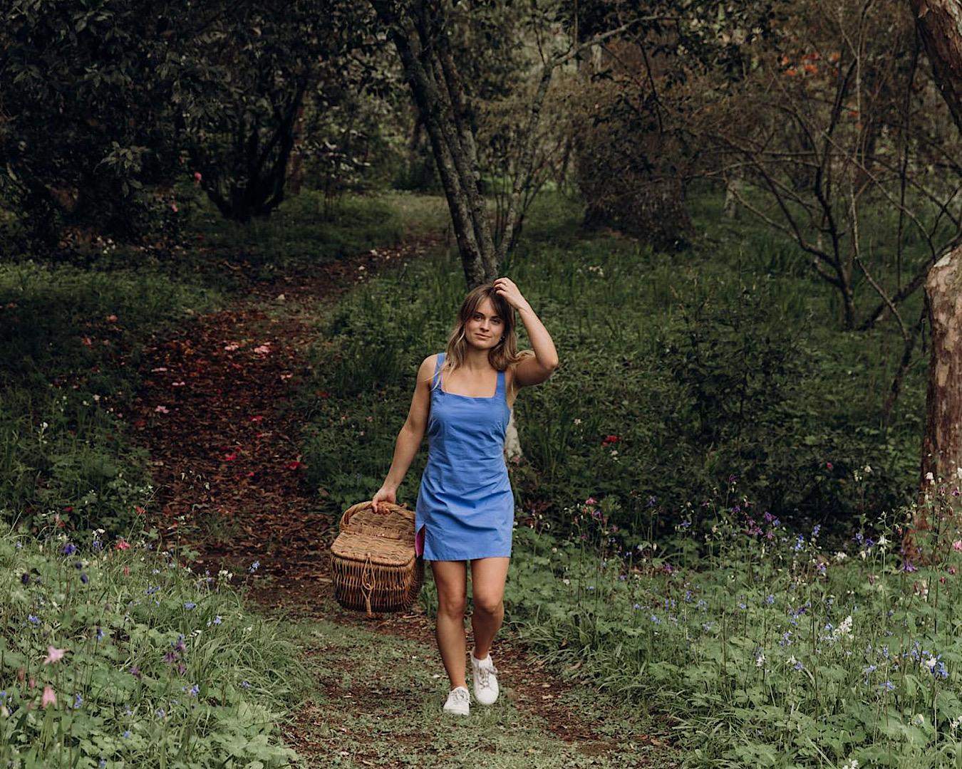 A woman in a reversible cornflower blue-pink dress walks through a forest carrying a picnic basket. 