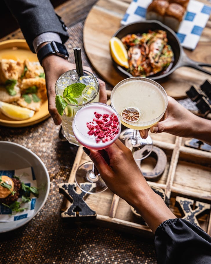 Beautiful cocktails clink above a table filled with gorgeous food. 