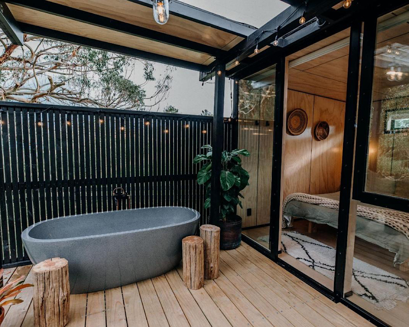 A large stone bathtub sits on a deck under a string of dolly lights. Inside, we see the edge of a cosy bed. 