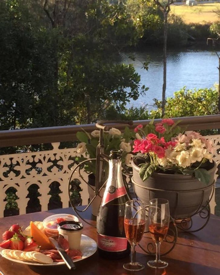Shows golden hour light hitting a table on a front deck, overlooking a river, surrounding greenery and a sunny paddock opposite. On the table there is a bottle of nice champagne, two full glasses next to a charcuterie plate and a pot of pink and white flowers. .