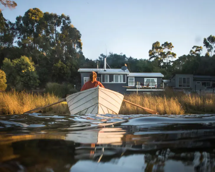 A person rows a boat in front of Blythe River Boathouse lake front Airbnb Tasmania