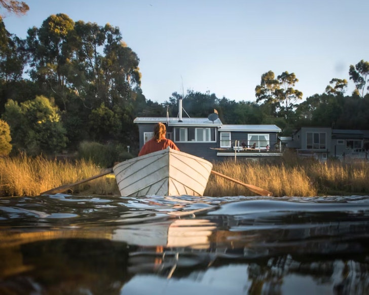 A person rows a boat in front of Blythe River Boathouse lake front Airbnb Tasmania